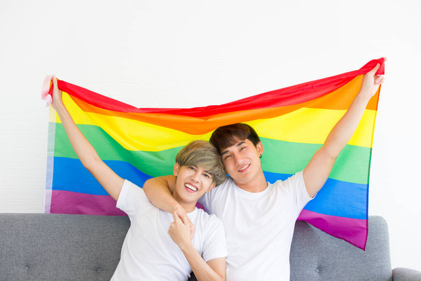 Gelukkig Aziatische homoseksuele mannen of homoseksuele stellen omarmen elkaar samen met een regenboogvlag zittend op de bank. Concept van LGBTQ-trots. - Foto, afbeelding