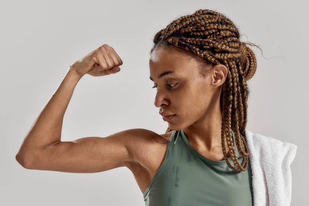 Get stronger. Close up portrait of young sportive mixed race woman showing her biceps after intensive training isolated over grey background - Photo, Image