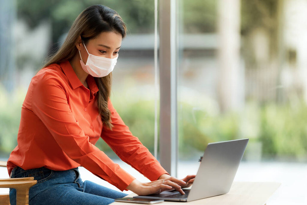 Portrait of young woman wearing face mask protective using a laptop sitting near window in creative office or cafe - Foto, Bild