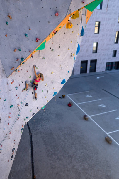 Μια γυναίκα σκαρφαλώνει σε τοίχο αναρρίχησης, ένας ορειβάτης εκπαιδεύεται σε τεχνητό έδαφος, αναρρίχηση στην πόλη, ένα δυνατό κορίτσι, σπορ στην πόλη, ασφάλεια στα ακραία αθλήματα. - Φωτογραφία, εικόνα