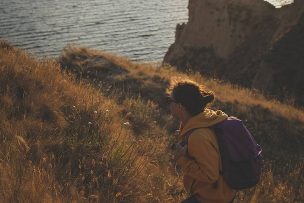 Young woman hiker in yellow hoodie with backpack climbing rock on mountain peak cliff by the ocean during autumn sunset. Soft filter. Adventure wanderlust active lifestyle concept.  - Photo, Image