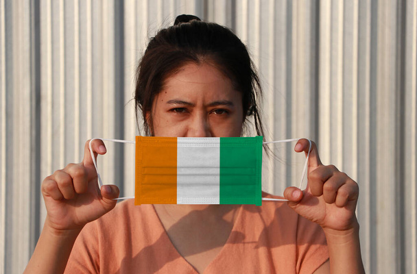 Μια γυναίκα με Ivory Coat σημαία σε υγιεινή μάσκα στο χέρι της και σήκωσε το μπροστινό πρόσωπο σε γκρι φόντο. Μικροσκοπική προστασία μορίων ή ιών ή Covid 19. Έννοια της καταπολέμησης της ασθένειας. - Φωτογραφία, εικόνα