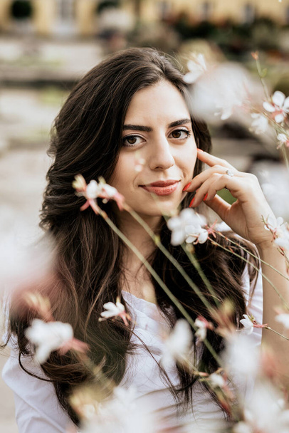 Κάθετη φωτογραφία χαμογελαστή νεαρή γυναίκα που περιβάλλεται από λουλούδια υπαίθρια. Μελαχρινή γυναίκα σε φόντο φρέσκα λουλούδια - Φωτογραφία, εικόνα