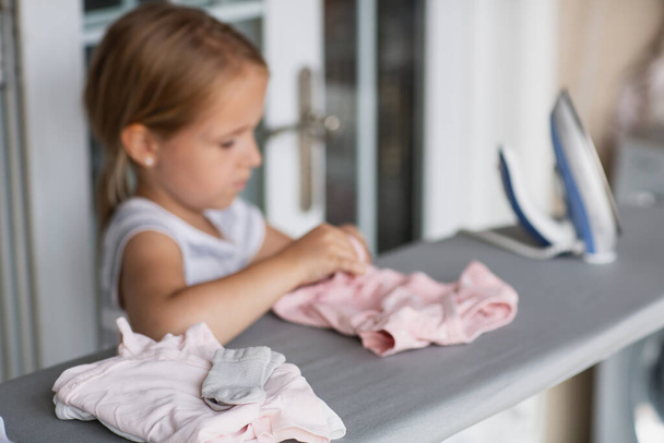 Cute little girl with blonde hair is leaning on ironing clothes on board at home. High quality photo - Valokuva, kuva