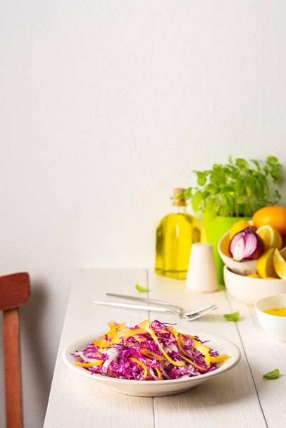 Lehký letní salát z červeného zelí, cibule a žluté papriky, salát na talíři na bílém dřevěném stole, olivový olej s kořením na dresink a bazalku s koprem - Fotografie, Obrázek