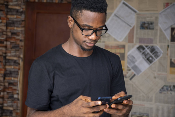 Une mise au point peu profonde d'un jeune homme africain avec des lunettes utilisant deux téléphones dans une pièce - Photo, image