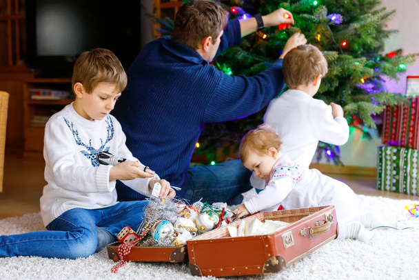 Két kisfiú és imádnivaló kislány, akik karácsonyfát díszítenek régi játékokkal és labdákkal. Apa a háttérben - Fotó, kép