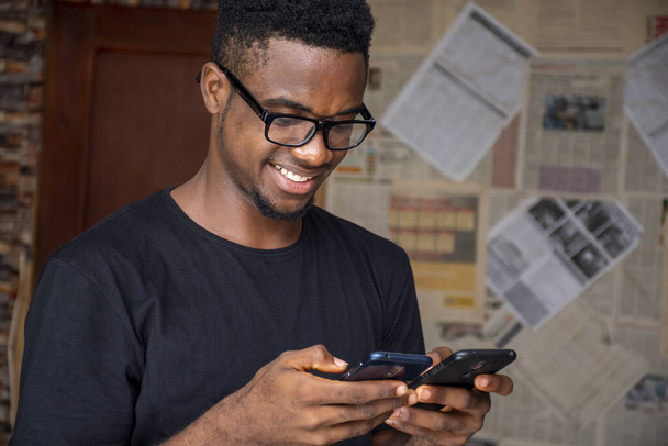 A shallow focus of a young African male with glasses using two phones in a room - 写真・画像