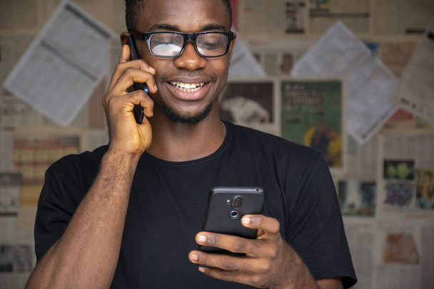 A young African male with glasses talking on the phone while using another one in a room - 写真・画像