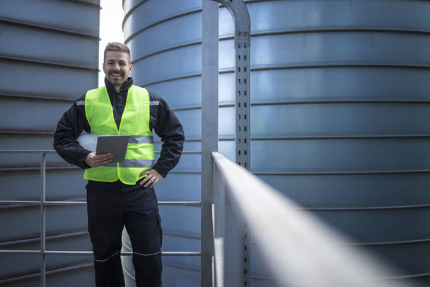 Portrait of factory engineer worker standing on metal platform between industrial storage tanks and looking to the camera. - Fotografie, Obrázek