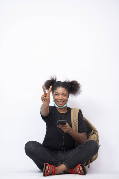 A pretty African female carrying a backpack, sitting legs crossed using her mobile phone, making the peace sign - Foto, afbeelding