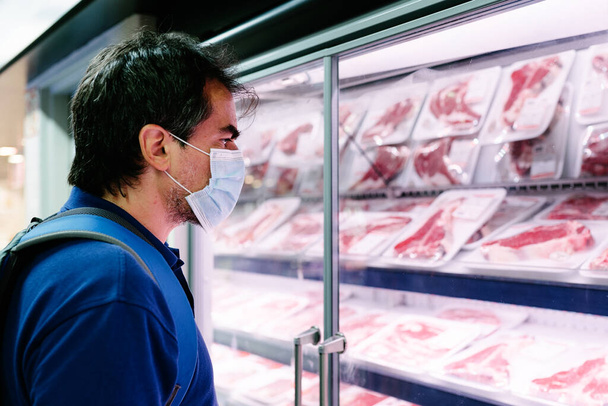 Un turista adulto de mediana edad con una mascarilla higiénica de compras en la sección de carne refrigerada de un supermercado - Foto, Imagen