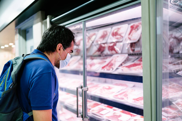 Um turista adulto de meia-idade com uma máscara higiênica compras na seção de carne refrigerada de um supermercado - Foto, Imagem