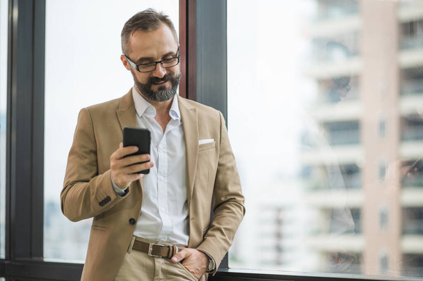 Молодой красивый бизнес бородатый мужчина стоит у окна, используя смартфон на рабочем месте офиса. - Фото, изображение