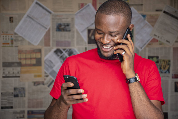 Un giovane maschio africano con gli occhiali che parla al telefono mentre ne usa un altro in una stanza - Foto, immagini