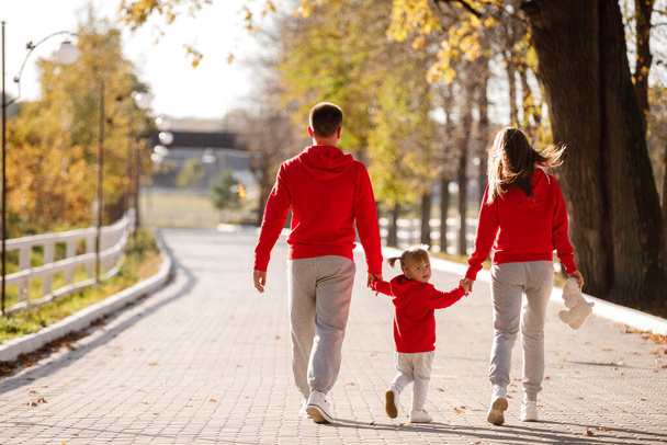 Мама, папа держатся за руки дочери и гуляют в осеннем парке. Молодая семья проводит время вместе на отдыхе, на открытом воздухе. Концепция семейного отдыха - Фото, изображение