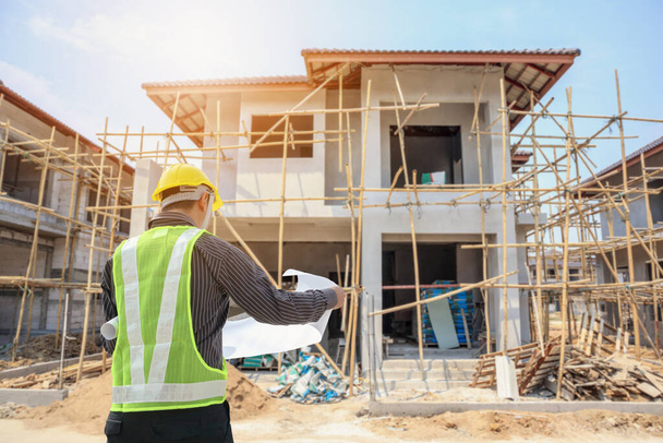 Профессиональный инженер-архитектор с защитным шлемом и чертежной бумагой на фоне строительства дома - Фото, изображение