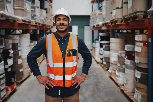 Portrait of successful manager standing in warehouse between shelf filled with goods wearing a white helmet and orange vest for protection with hands on waist - Фото, изображение