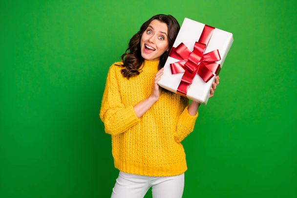 Photo of funny pretty lady hold big paper box red bow near head ear birthday party event gift wear knitted yellow sweater white trousers isolated vibrant green color background - Фото, изображение