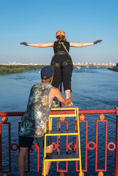 Nierozpoznawalna odważna dziewczyna przygotowująca się do skoku na bungee z mostu przez rzekę przy wsparciu instruktora. Widok z tyłu. Koncepcja sportów ekstremalnych. Orientacja pionowa - Zdjęcie, obraz