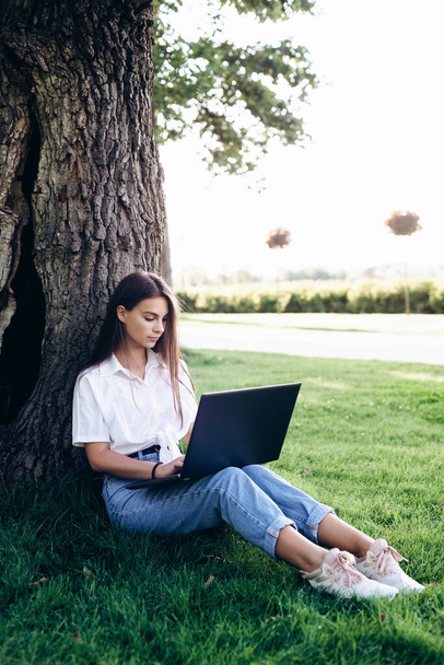 Tyttö opiskelija kannettava tietokone ulkona istuu nurmikolla puistossa lähellä puuta, surffailu internetissä tai valmistautuu tentit. Teknologia, koulutus ja etätyön käsite. Pehmeä valikoiva kohdentaminen. - Valokuva, kuva