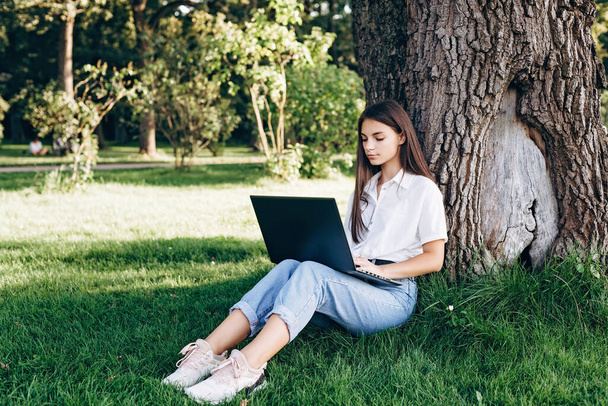 студентка з ноутбуком на відкритому повітрі сидить на траві в парку біля дерева, займається серфінгом в Інтернеті або готується до іспитів. Технології, освіта та концепція дистанційної роботи. М'який вибірковий фокус
. - Фото, зображення