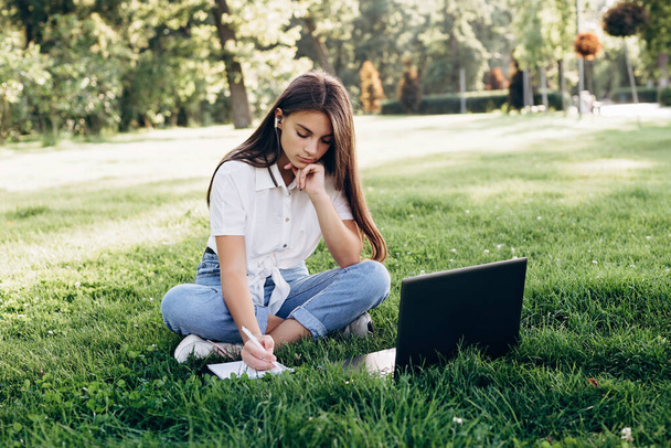 beautiful young girl student with a laptop outdoors sitting on the grass, surfing the internet or preparing for exams. Technology, education and remote work concept. Soft selective focus. - Photo, Image