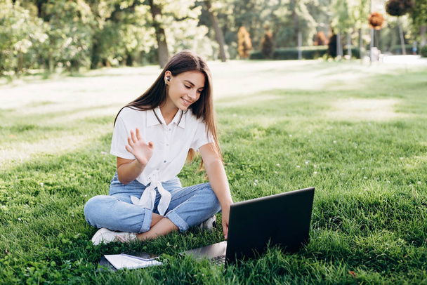ノートパソコンを持った女子大生。笑顔の女性は、コンピュータで芝生の上に座って、インターネットサーフィンや試験の準備。技術、教育、遠隔作業の概念。柔らかい選択的フォーカス. - 写真・画像