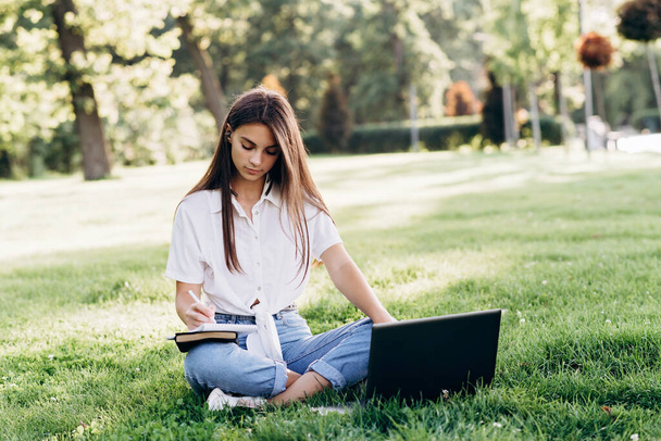 gyönyörű fiatal lány diák laptoppal a szabadban ül a füvön, felkészülve a vizsgákra. Technológia, oktatás és távmunka. Puha szelektív fókusz. - Fotó, kép