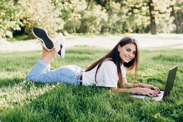 student girl with laptop outdoors. Smiling woman lying on the grass with a computer, surfing the Internet or preparing for exams. Technology, education and remote work concept. Soft selective focus. - Фото, зображення
