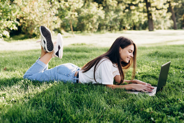 student girl with laptop outdoors. Smiling woman lying on the grass with a computer, surfing the Internet or preparing for exams. Technology, education and remote work concept. Soft selective focus. - Photo, Image