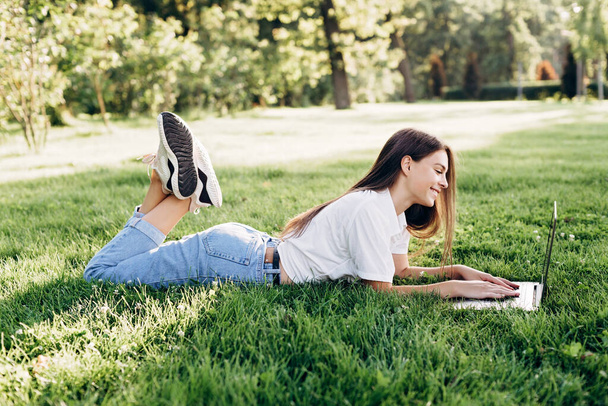 student girl with laptop outdoors. Smiling woman lying on the grass with a computer, surfing the Internet or preparing for exams. Technology, education and remote work concept. Soft selective focus. - Фото, зображення