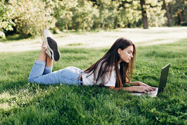 student girl with laptop outdoors. woman lying on the grass with a computer, surfing the Internet or preparing for exams. Technology, education and remote work concept. Soft selective focus. - Photo, image