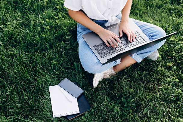 屋外にノートパソコンを持っている学生の女の子は、芝生の上に座って、インターネットをサーフィンや試験の準備。技術、教育、遠隔作業の概念。柔らかい選択的フォーカス. - 写真・画像