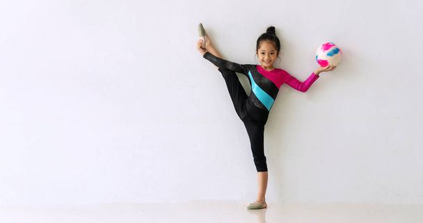 Acrobat mała słodka dziewczyna stojąca jedną nogę trzymając kolorowy piłkę w ręce w pokoju na białym tle. Urocza dziewczynka robi gimnastykę balansować swoje ciało na podłodze. Koncepcja ćwiczeń. - Zdjęcie, obraz