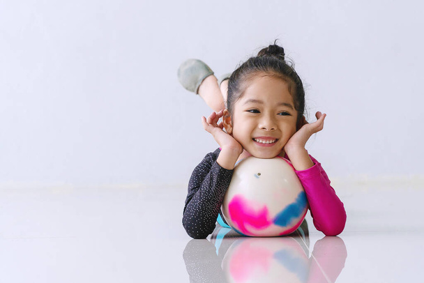 Liebenswertes kleines Mädchen, das mit Ball auf dem Boden liegt und in die Kamera schaut. Fröhliches Kind entspannt sich nach der Gymnastik im Studio. Wellness-Lifestyle-Konzept. - Foto, Bild