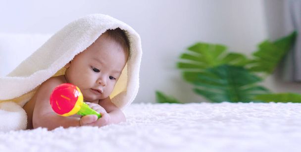 Veselé roztomilé dítě ležící na posteli s hračkou pod bílou přikrývkou a dívající se na něco. Nevinný dítě plazí na bílé posteli s ručníkem na hlavě doma a drží hračku v ruce. - Fotografie, Obrázek