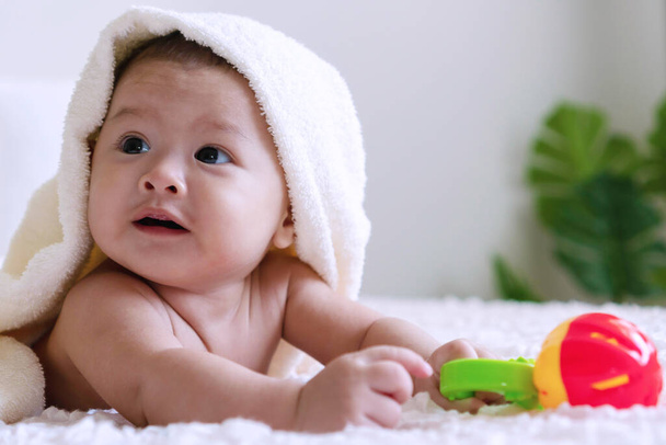 Vrolijke schattige baby liggend op bed met speelgoed onder witte deken op zoek naar iets. Onschuld baby kruipend op wit bed met handdoek op zijn hoofd thuis en met speelgoed in zijn hand. - Foto, afbeelding