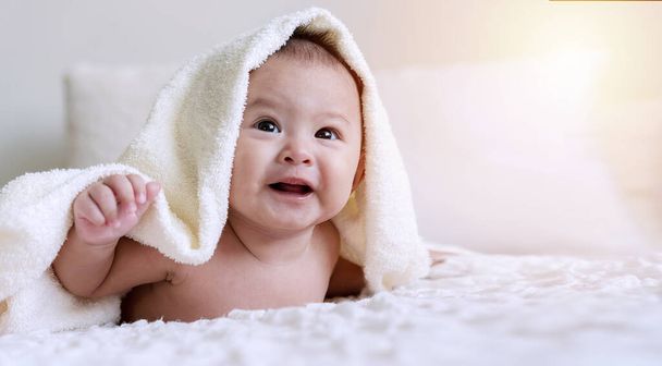 Χαρούμενο χαριτωμένο μωρό που κοιτάζει την κάμερα κάτω από λευκή κουβέρτα κοιτάζοντας κάτι. Αθώο μωρό σέρνεται στο λευκό κρεβάτι με πετσέτα στο κεφάλι του στο σπίτι. - Φωτογραφία, εικόνα
