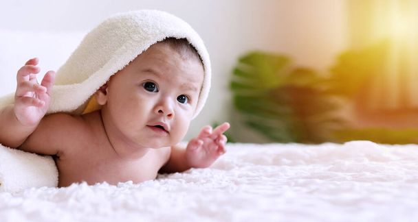 Fröhliches süßes Baby, das unter einer weißen Decke in die Kamera schaut und etwas ansieht. Unschuld Baby kriecht auf weißem Bett mit Handtuch auf dem Kopf zu Hause. - Foto, Bild