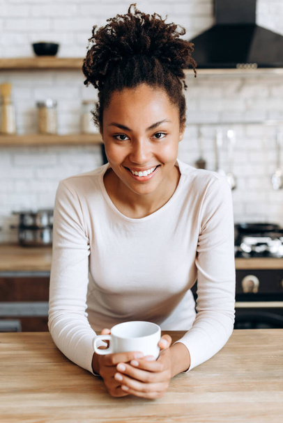 Gyönyörű nő alkalmi ruhákban egy csészével a kezében, a kamerába néz és mosolyog, miközben a konyhában ül. - Fotó, kép