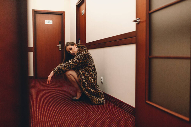 A young beautiful girl in the evening at the hotel alone walks along the corridor between the rooms. she's wearing black underwear and a spotted fur coat - Foto, afbeelding