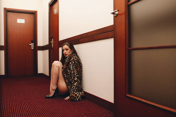 Ein junges, schönes Mädchen läuft abends allein im Hotel den Gang zwischen den Zimmern entlang. Sie trägt schwarze Unterwäsche und einen gefleckten Pelzmantel - Foto, Bild