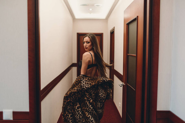 A young beautiful girl in the evening at the hotel alone walks along the corridor between the rooms. she's wearing black underwear and a spotted fur coat - 写真・画像