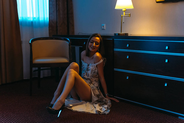 A young beautiful girl in the evening at the hotel alone, sits on the floor. Evening light from Sconces - Photo, image
