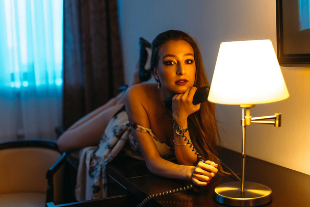 A young beautiful girl is alone at the hotel in the evening, sitting at the table and talking on the phone. Evening light from Sconces - Foto, Imagem