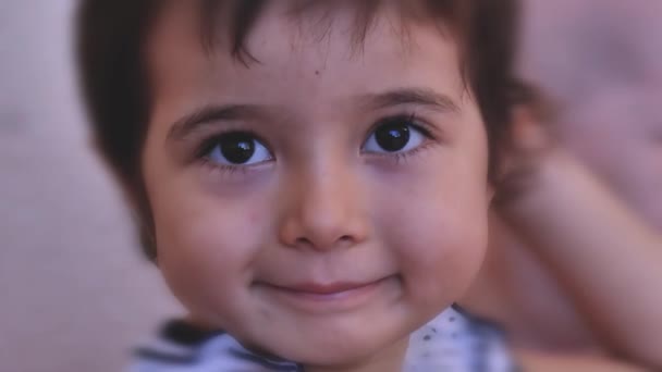 子供は笑顔でカメラを覗き込む。表情豊かな瞳の少女が微笑む。子孫の顔の一部がぼんやりとその側面に表示されます。閉鎖. - 映像、動画