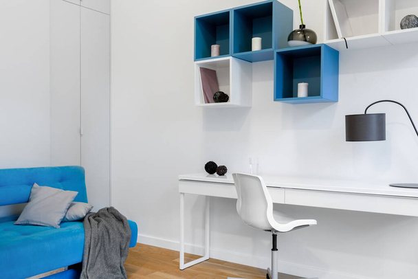 Jednoduchý a moderní studentský pokoj s bílým stolem, modrým gaučem a moderními čtvercovými policemi - Fotografie, Obrázek