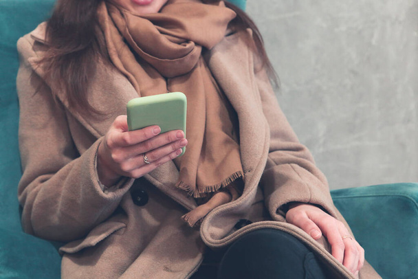 Crop female in warm brown coat and scarf interacting with smartphone while sitting in soft armchair in corridor of contemporary high rise building - Foto, Imagem