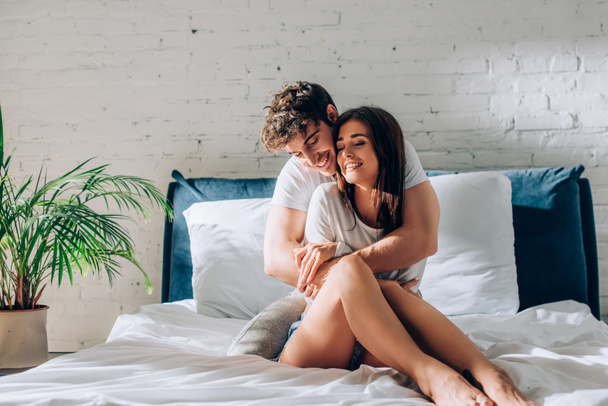 Jovem casal com os olhos fechados sentados na cama e abraçando uns aos outros - Foto, Imagem
